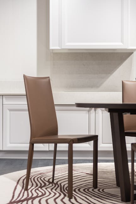 A close-up view of a modern dining area featuring a brown leather-upholstered chair with sleek, slim legs positioned next to a dark wooden table. The area has a neutral color palette, with a light bei