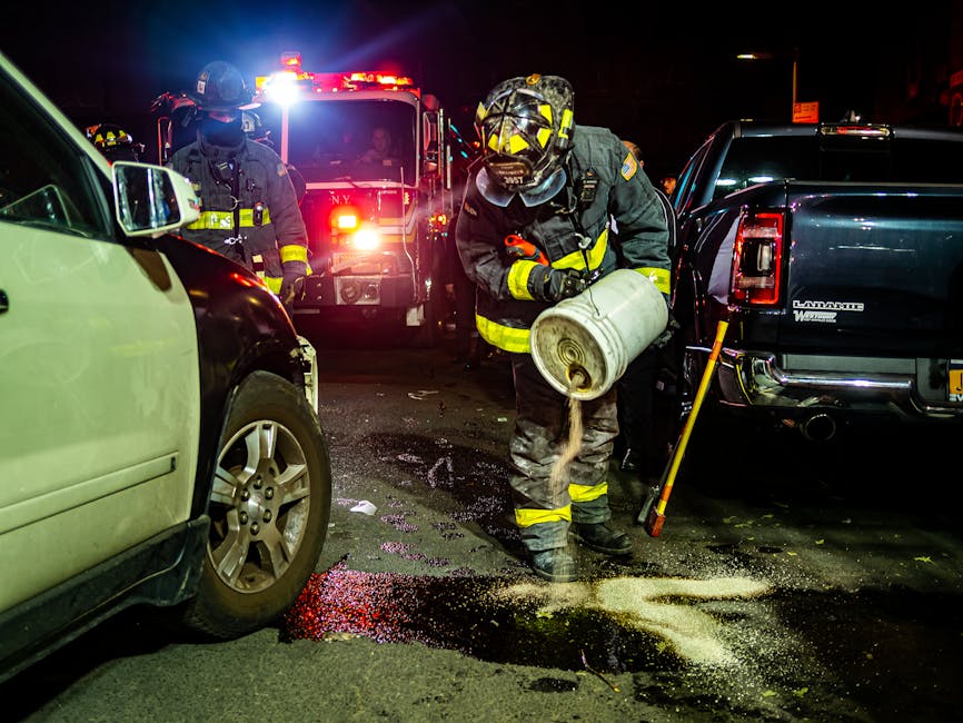 Nighttime scene of firefighters in full protective gear and helmets attending to an emergency spill on a city street near Bond Street, Mayfair. The firefighter in the foreground is pouring a granular substance from a white container onto the wet asphalt, likely for decontamination or spill containment. Bright lights from emergency vehicles, including a fire truck, illuminate the background, casting reflections on nearby vehicles, including a green and a black car parked closely on the street. The scene depicts active surface cleaning and hazard management as part of emergency response efforts, with the street surface appearing wet and partially covered with the spilled material. The area is illuminated by streetlights and emergency lighting, emphasizing the critical moment of spill cleanup coordinated by Mayfair Carpet Cleaners and emergency services.