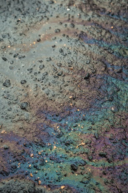 Close-up view of a wet, textured concrete or stone surface with scattered small pebbles and debris, displaying a mix of dark grey, brown, and iridescent blue-green hues from surface moisture or cleaning residue. The surface appears to have been recently exposed to a spill or cleaning process, with visible wet patches and uneven texture. The surrounding area suggests an outdoor or industrial environment, with natural lighting highlighting the roughness and dampness of the material. Mayfair Carpet Cleaners in Mayfair provides expert surface cleaning and sanitisation services, ensuring thorough removal of contaminants from various surfaces.