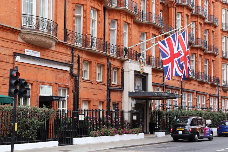 The exterior of Claridge's hotel in Mayfair, London, showcasing a classic red brick facade with multiple white-framed windows and black wrought iron balconies. Prominent Union Jack flags are displayed