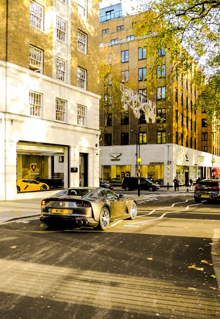 Street view of Mount Street W1K in Mayfair featuring a mix of commercial buildings with large display windows, some showcasing luxury cars like a yellow Lamborghini and a black sports car parked in front. The buildings are constructed from light-colored stone and dark brick materials, with multiple rectangular windows. The scene is illuminated by natural daylight, casting shadows from nearby trees with green and yellow leaves. The street surface is asphalt with visible road markings, and there are pedestrians walking along the sidewalk. Mayfair Carpet Cleaners logo and signage are not visible in this image, but the scene emphasizes the upscale urban environment that may require professional cleaning services for the commercial properties.