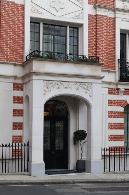 The exterior of a red brick residential building featuring white decorative stone accents around the arched entrance, which has a black door with glass panels and intricate patterns. Above the doorway, there is a large window with a black metal balcony railing decorated with greenery. The front area includes a small black iron fence, and a potted plant with a neatly trimmed bush is placed near the entrance. Natural daylight illuminates the facade, highlighting the clean and well-maintained appearance of the building. Mayfair Carpet Cleaners specializes in surface cleaning, deep cleaning, and sanitisation for properties like this, ensuring a pristine and hygienic environment for residents.