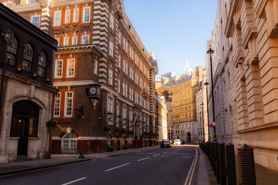 An urban street scene in Mayfair showing wet pavement and cobblestone sidewalks after rainfall, with reflections of nearby red-brick and cream-colored Victorian-style buildings featuring bay windows a